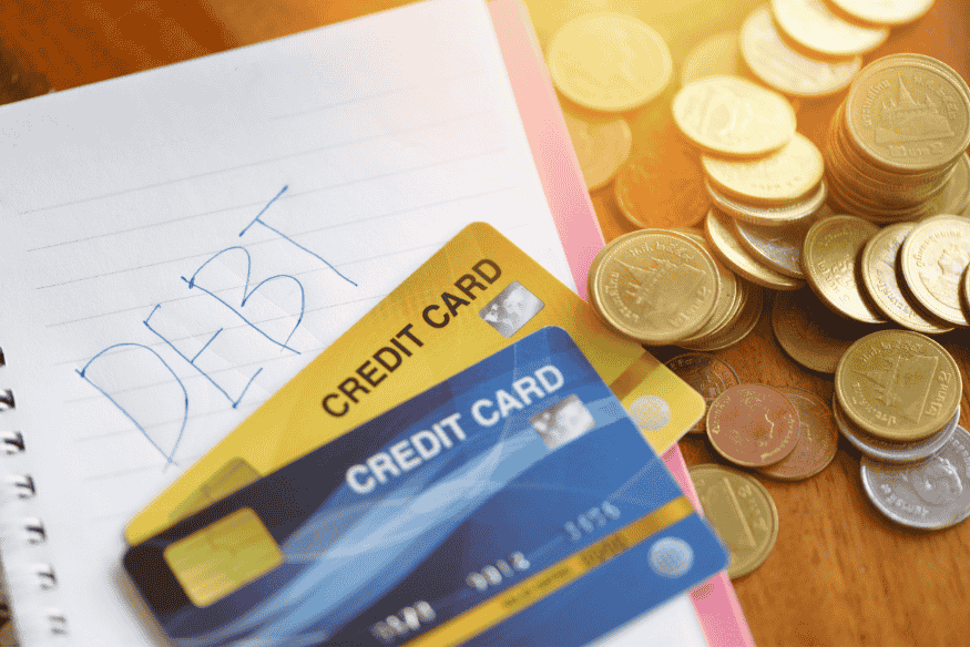 Credit cards on a notebook with the word "DEBT" written, next to piles of coins