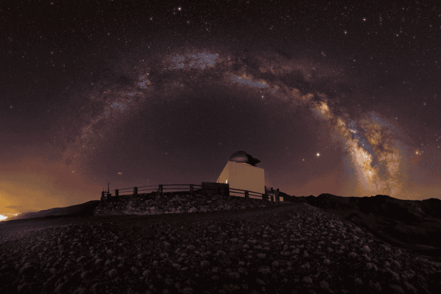 Via Láctea forma arco no céu estrelado sobre observatório astronômico em área remota.