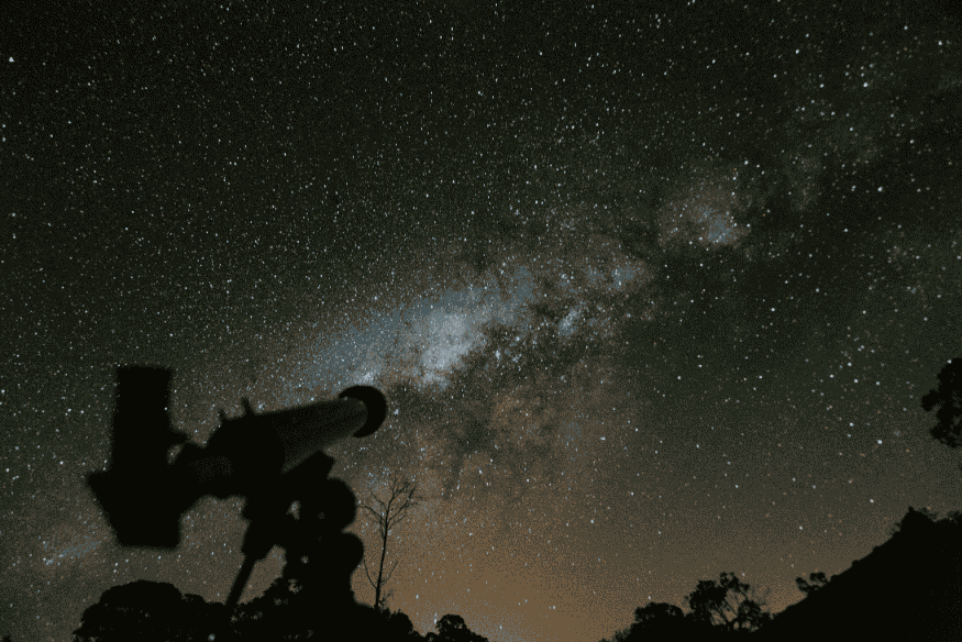 Silhueta de telescópio com Via Láctea visível ao fundo em céu escuro.