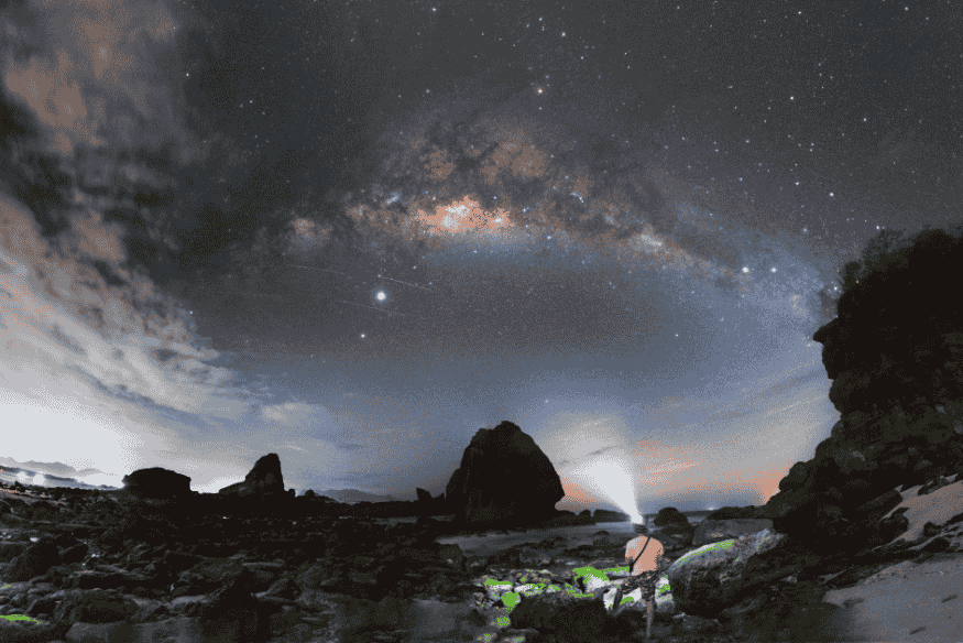 Via Láctea visível sobre formação rochosa, com pessoa iluminando o solo, ideal para astrofotografia de paisagem.