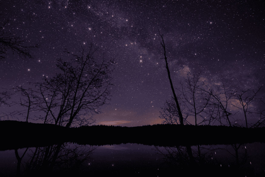Reflexo do céu estrelado em lago calmo, com silhuetas de árvores, capturado à noite.