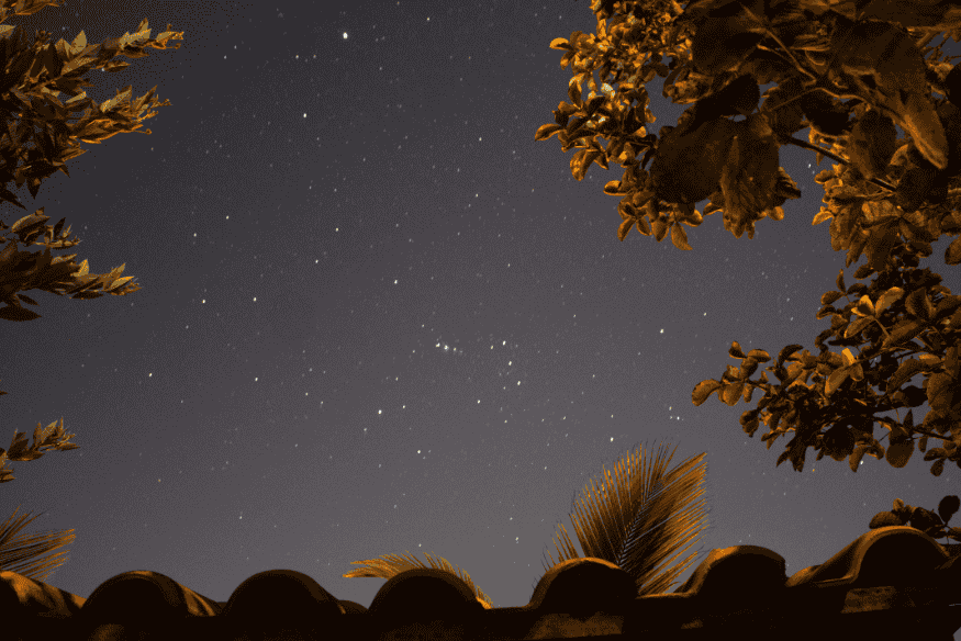 Céu noturno com estrelas visíveis em composição urbana, capturado com smartphone entre folhas e telhado.