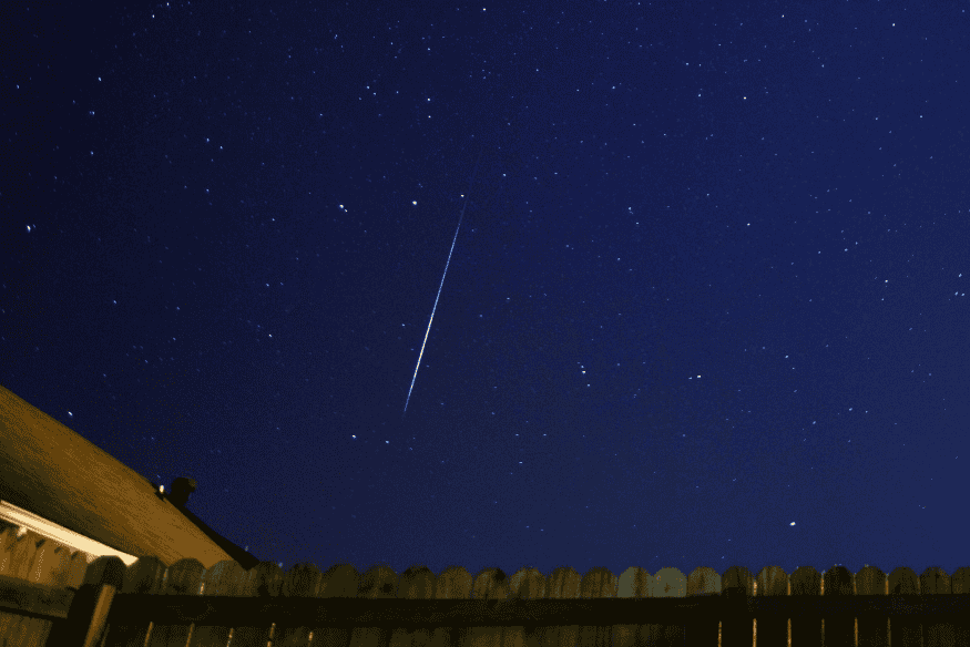 Rastro de meteoro visível no céu noturno estrelado acima de telhado e cerca residencial.