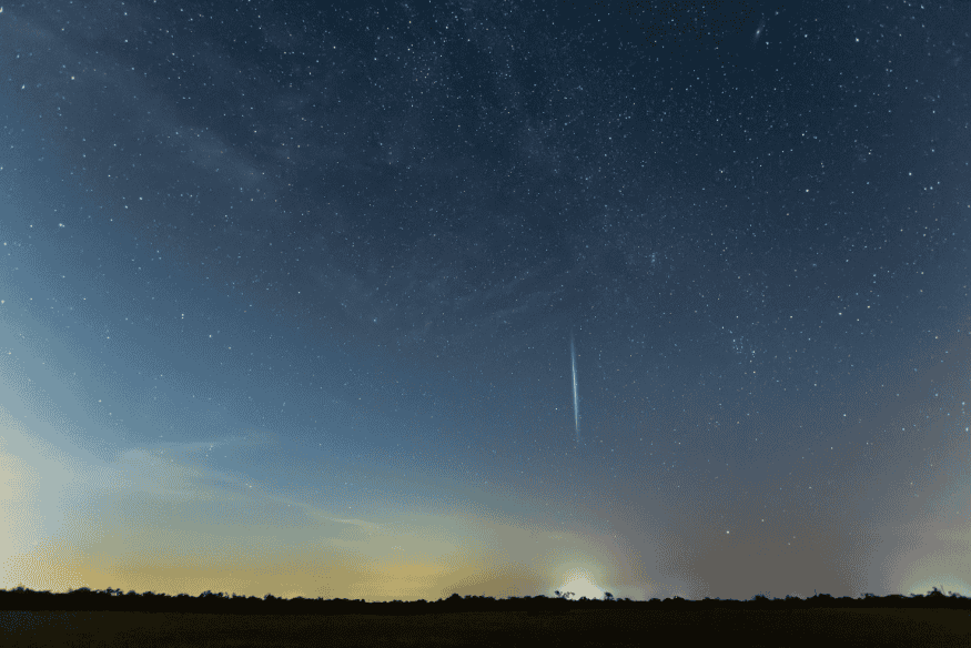 Meteoro cruzando o céu noturno sobre área rural com horizonte limpo e estrelas visíveis.