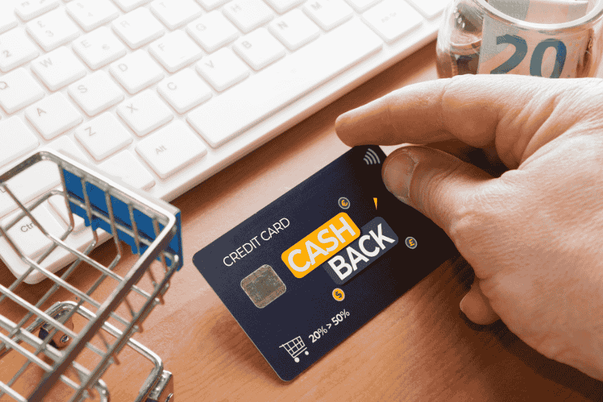Hand holding a cashback credit card near a keyboard with a mini shopping cart