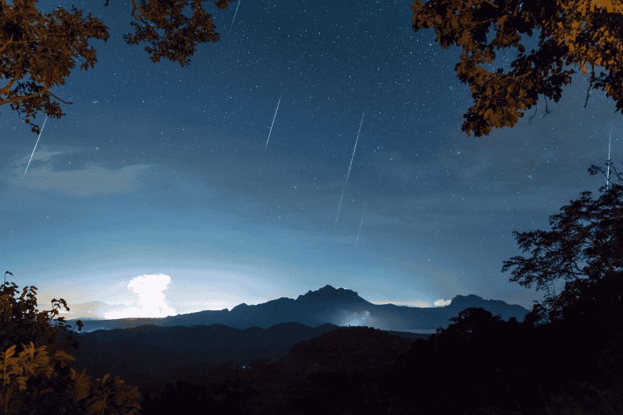 Céu limpo com estrelas e meteoros durante a chuva Gemínidas, sobre montanhas e árvores.