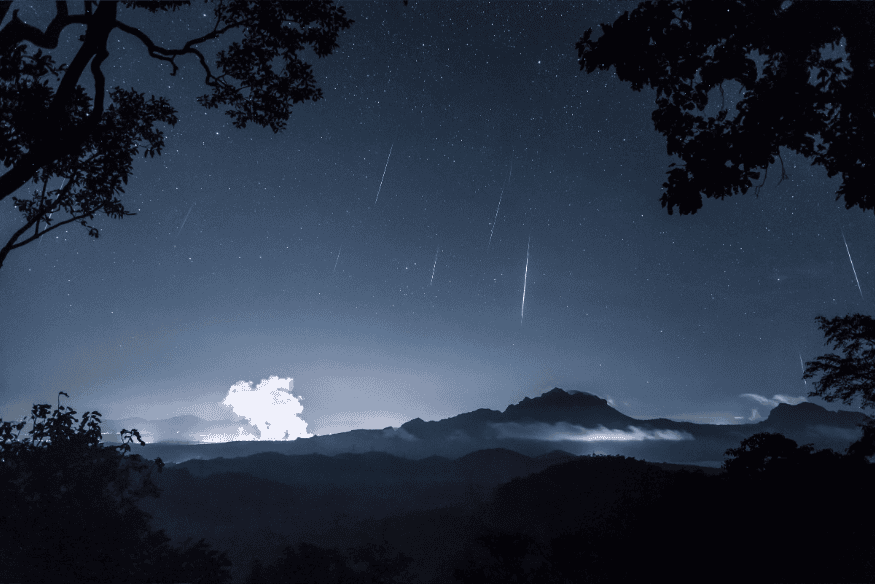 Trilhas de meteoros riscando o céu noturno sobre uma paisagem montanhosa com vegetação.