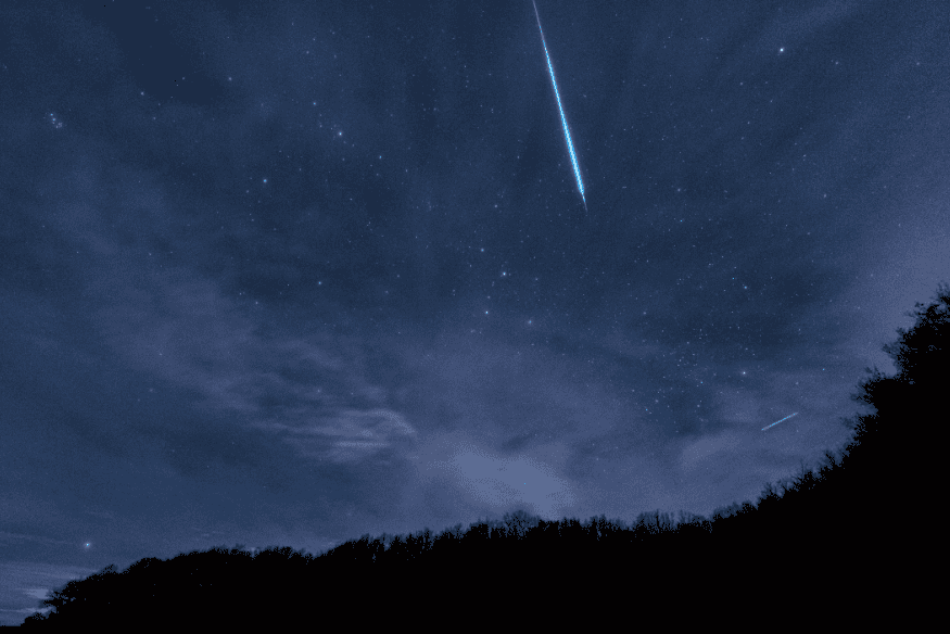 Meteoro riscando o céu estrelado sobre a silhueta de uma floresta à noite.