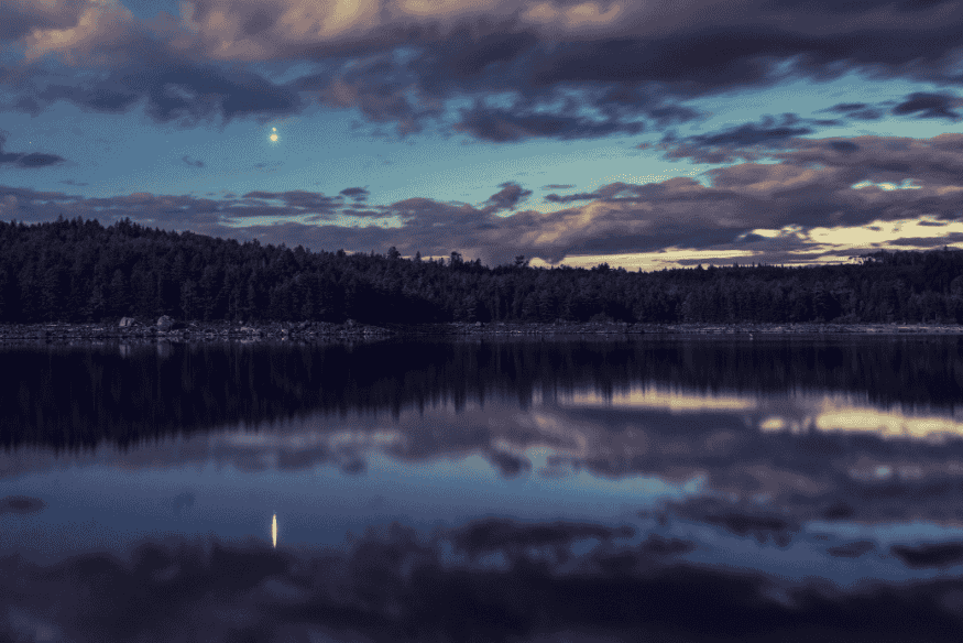 Planeta brilhante no céu ao anoitecer, com reflexo na água de um lago cercado por floresta.