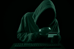 Hooded hacker inspecting a credit card under magnifying glass in a dark setting.