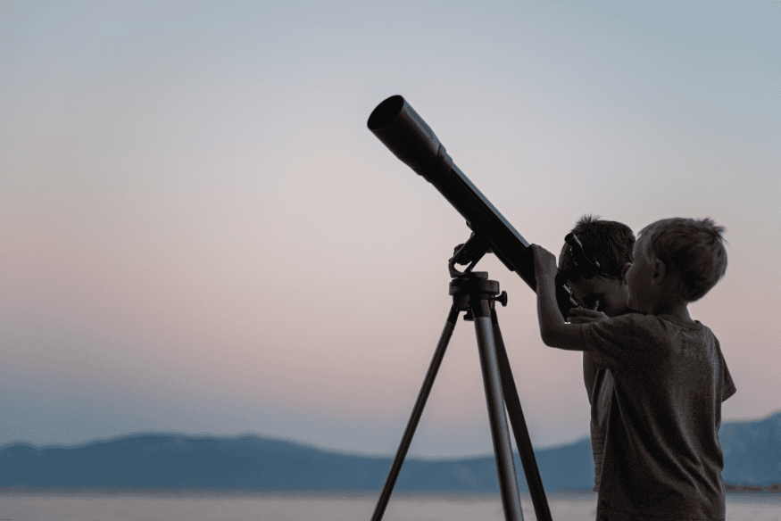 Duas crianças usam telescópio em ambiente externo durante o crepúsculo.