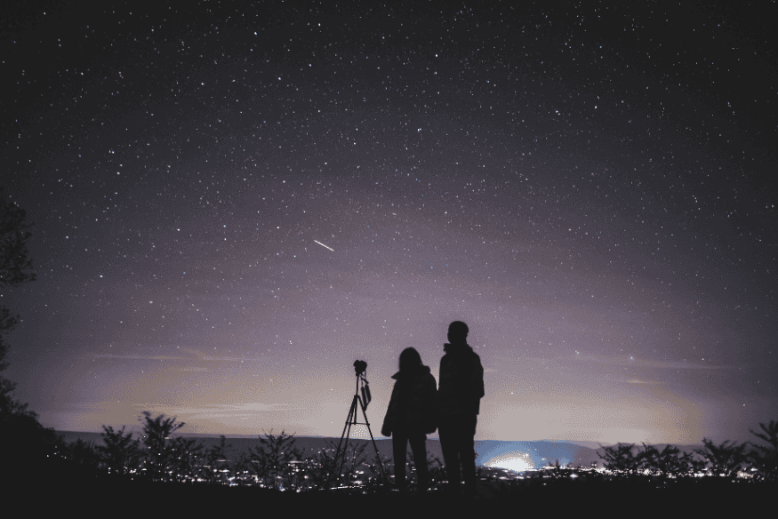 Casal observa o céu noturno estrelado com telescópio em local afastado da cidade.