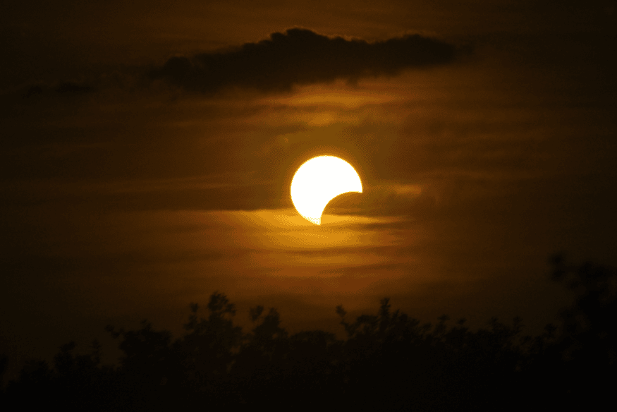 Eclipse solar parcial em céu alaranjado com nuvens e vegetação escura no horizonte.