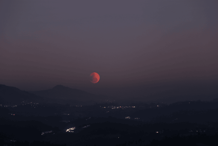 Eclipse lunar parcial com lua avermelhada surgindo no horizonte montanhoso ao anoitecer.
