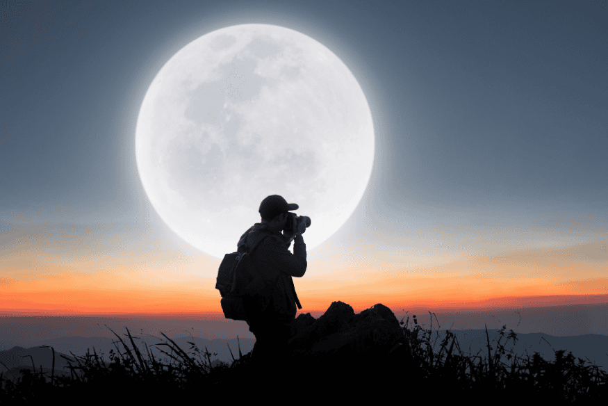 Pessoa fotografando a Lua cheia com câmera, tendo o astro ampliado ao fundo em pôr do sol.