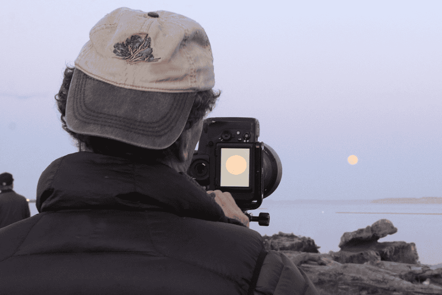 Tela de câmera mostrando a Lua enquanto fotógrafo ajusta o foco próximo a um lago.