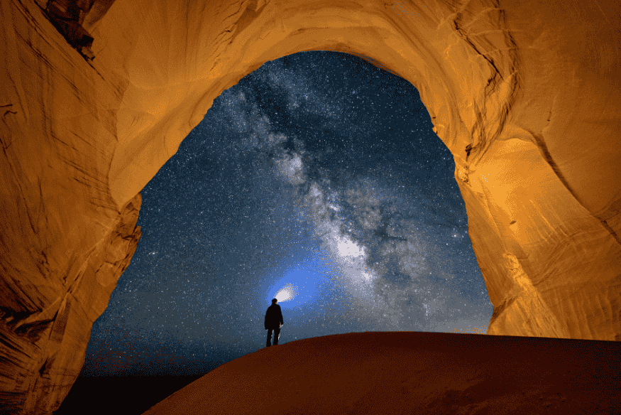 Pessoa com lanterna observa a Via Láctea através da entrada de uma caverna iluminada.