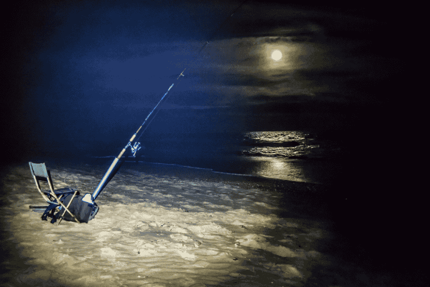 Vara de pesca fincada na areia durante a noite com lua cheia sobre o mar