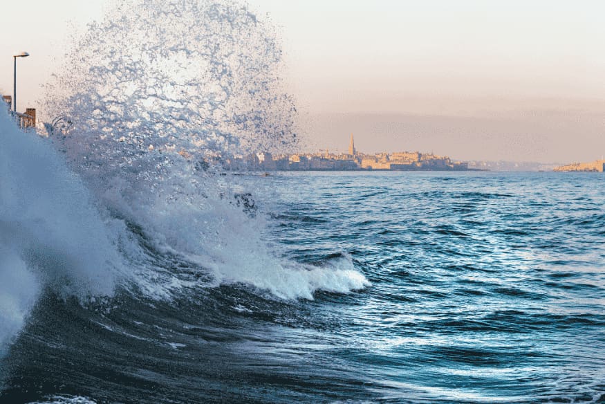 Grande onda se chocando contra o litoral com uma cidade ao fundo durante maré extrema.