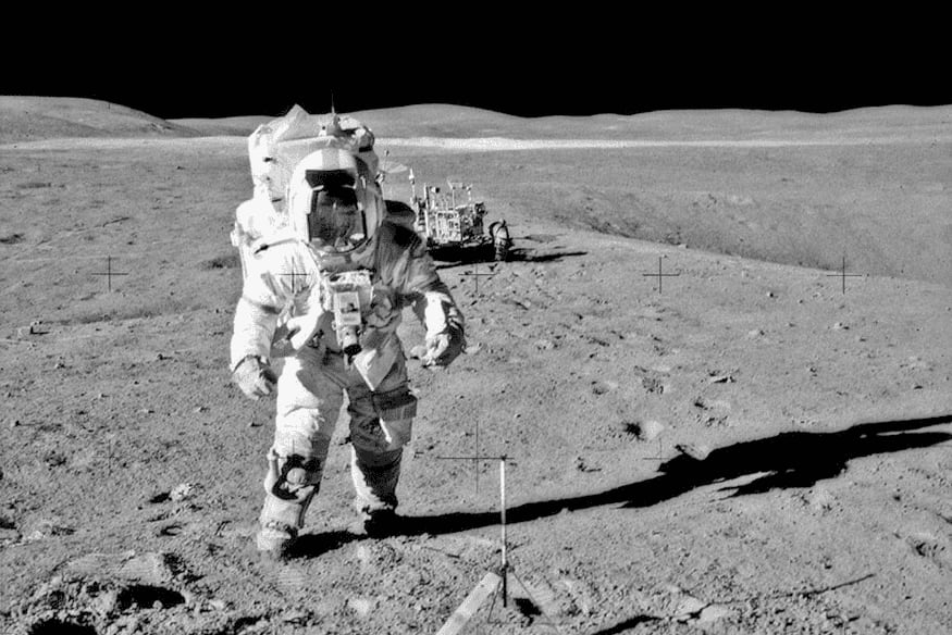 Astronauta caminhando na superfície lunar com traje espacial e veículo ao fundo durante missão Apollo.
