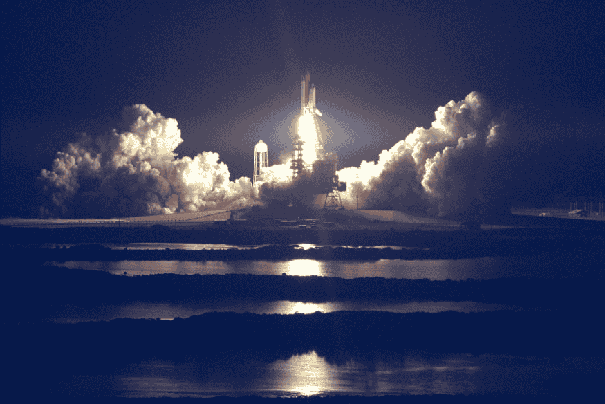 Lançamento de foguete espacial à noite com colunas de fumaça e luz refletida na água.