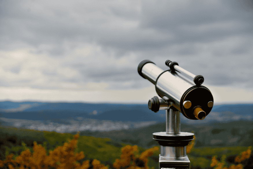 Telescópio metálico apontado para o céu em uma região montanhosa, com nuvens ao fundo