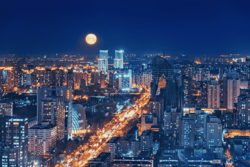 Vista noturna de cidade com iluminação intensa e lua cheia visível no horizonte.