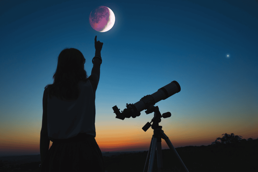 Mulher observando a Lua com telescópio ao entardecer, durante eclipse lunar parcial.