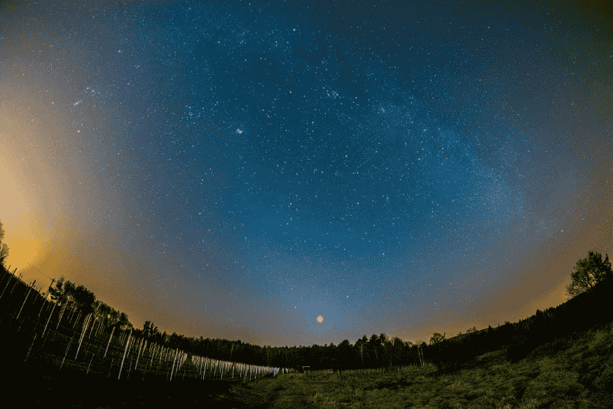 Campo aberto sob céu estrelado com lua e possíveis planetas visíveis no horizonte.