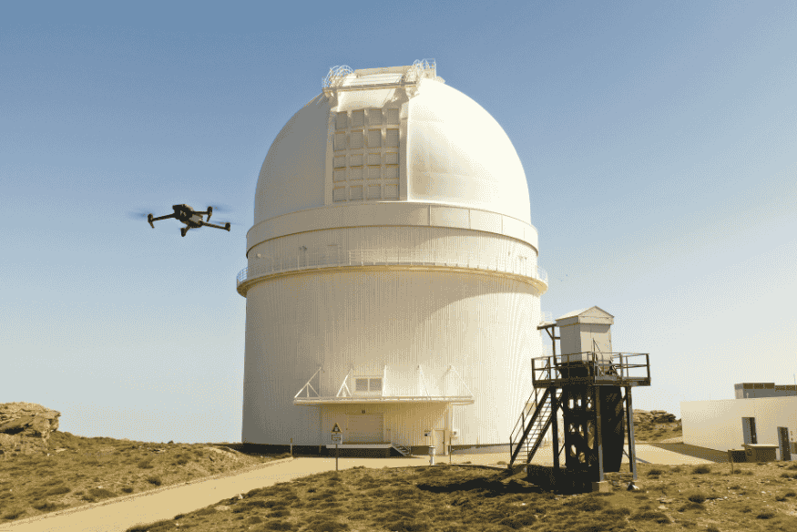 Observatório moderno com cúpula branca e drone sobrevoando instalações científicas em área remota.