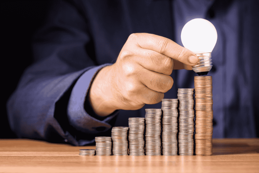 Hand placing a lit lightbulb on top of stacked coins, symbolizing financial ideas and investment growth