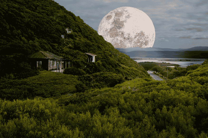 Superlua surgindo no horizonte atrás de colinas e vegetação, compondo uma paisagem natural.