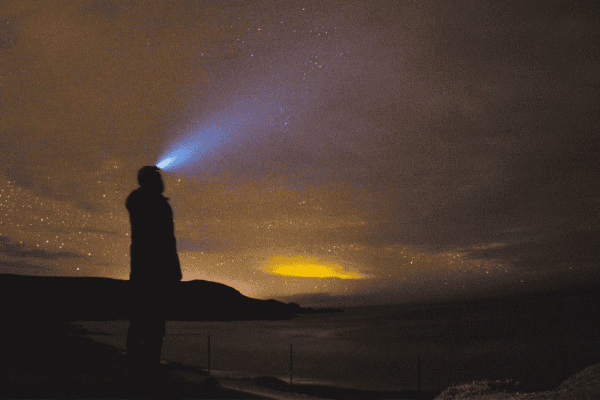 Viajante usando lanterna de cabeça enquanto observa o céu estrelado à beira-mar.